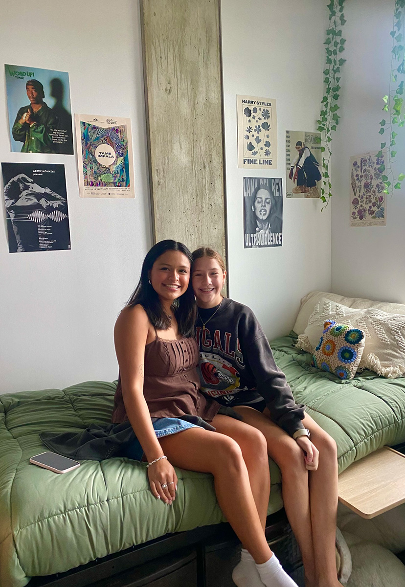 Roommates sitting on bed with green comforter, vines on wall