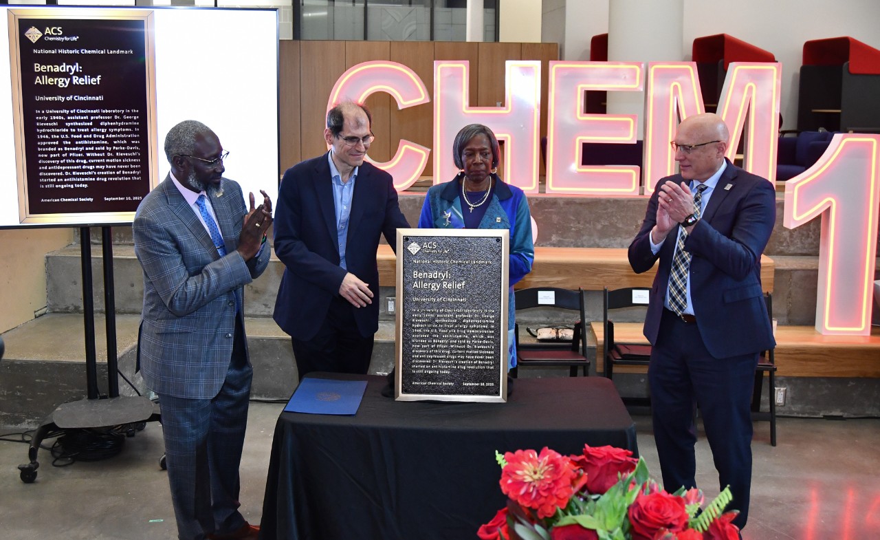 Four people unveil a plaque in front of a neon sign reading Chem 150.