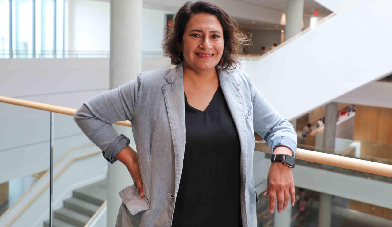 A woman in a gray jacket and black shirt leans against a railing.